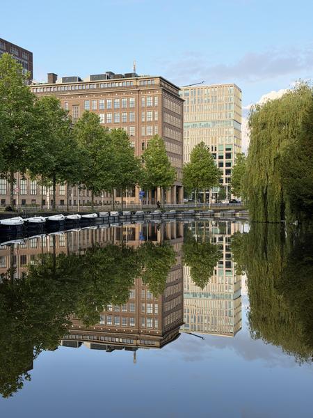 This tranquil photo captures a picturesque canal scene at Mauritskade, Oost, Amsterdam, showcasing the calm water perfectly reflecting the surrounding buildings and lush green trees
